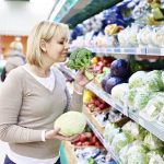 Mujer en el supermercado con una col en la mano, huele un brócoli delante del expositor de verdura repleto de coles