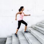 mujer morena vestida con malla pirata negra y camiseta amplia de deporte sin mangas subiendo escaleras de cemento gris
