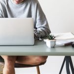 Mesa de trabajo con ordenador portatil, una plantita pequeña, un smartphone negro, y diversos cuadernos. Una mujer está sentada con las piernas desnudas cruzadas. Lleva un jersey gris y sonríe mientras mira la pantalla del portátil.