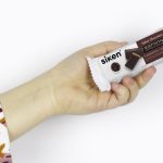 Primer plano de brazo con blusa blanca con flores marrones sujeta barrita sustitutiva de cacao Siken