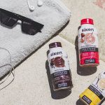 batidos sustitutivos Siken cacao, fresa yogur y vainilla sobre el suelo junto a una tolla, con unas gafas de sol y unos auriculares para escuchar música